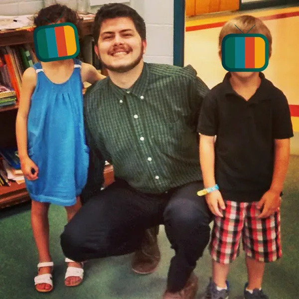 David student teaching at twenty-two, kneeling between two young students whose faces are covered by the site's television logo.